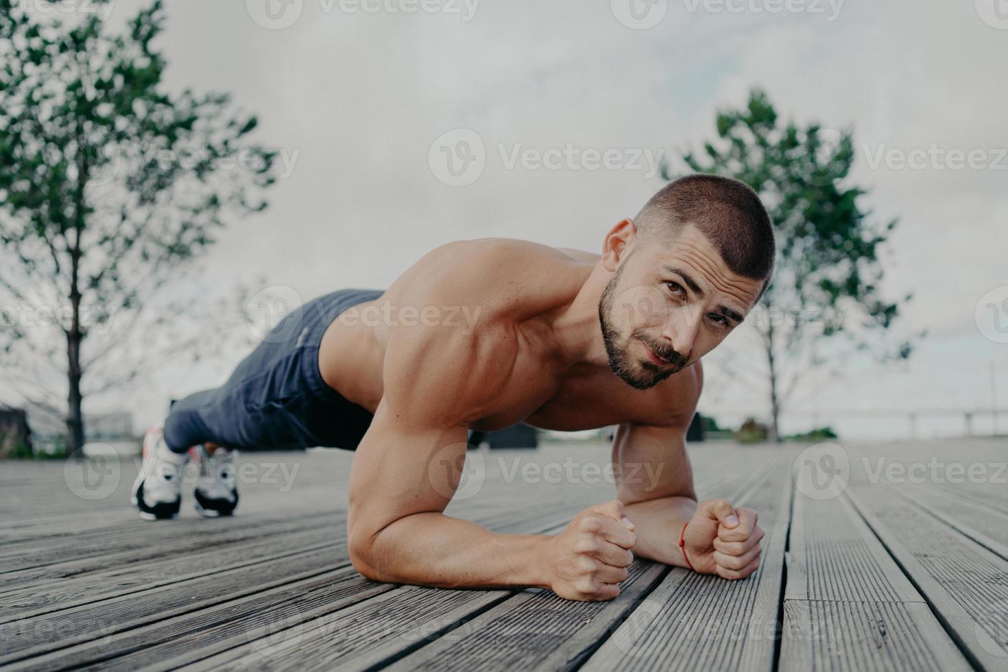 Motivational bearded man stands in plank pose, trains muscles and has strong body. Sporty adult guy performs push up exercise outdoor. Bodybuilder has abdominal workout. Active lifestyle concept photo