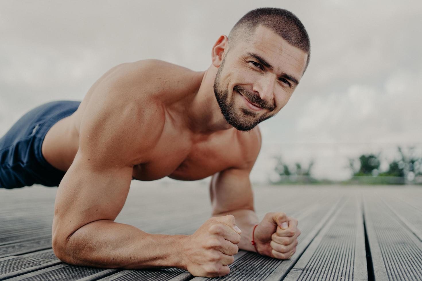 guapo deportista barbudo se para en tablón y practica deporte al aire libre, tiene entrenamiento regular y trabaja en los músculos, demuestra sus bíceps, trata de hacer lo mejor que puede. el hombre empuja hacia arriba el ejercicio. foto