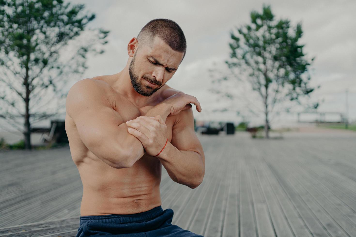 Outdoor shot of muscular guy injured elbow during physical exercises, feels strong pain and poses with naked torso. Health problems, medical, sickness and sport concept. Pain relief. Muscle injury photo