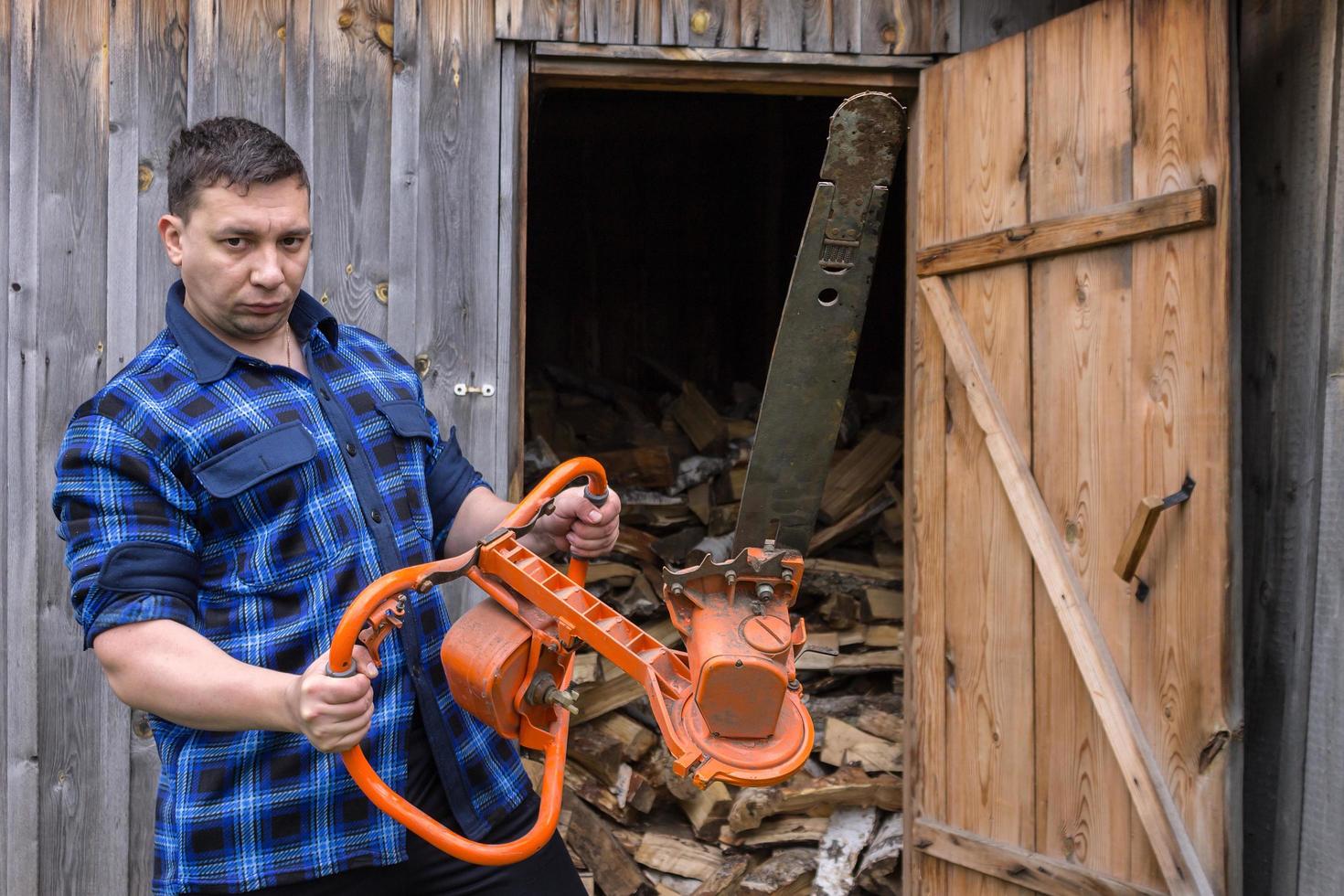 Farmer in a blue shirt with a big chainsaw on a background of a wooden warehouse photo