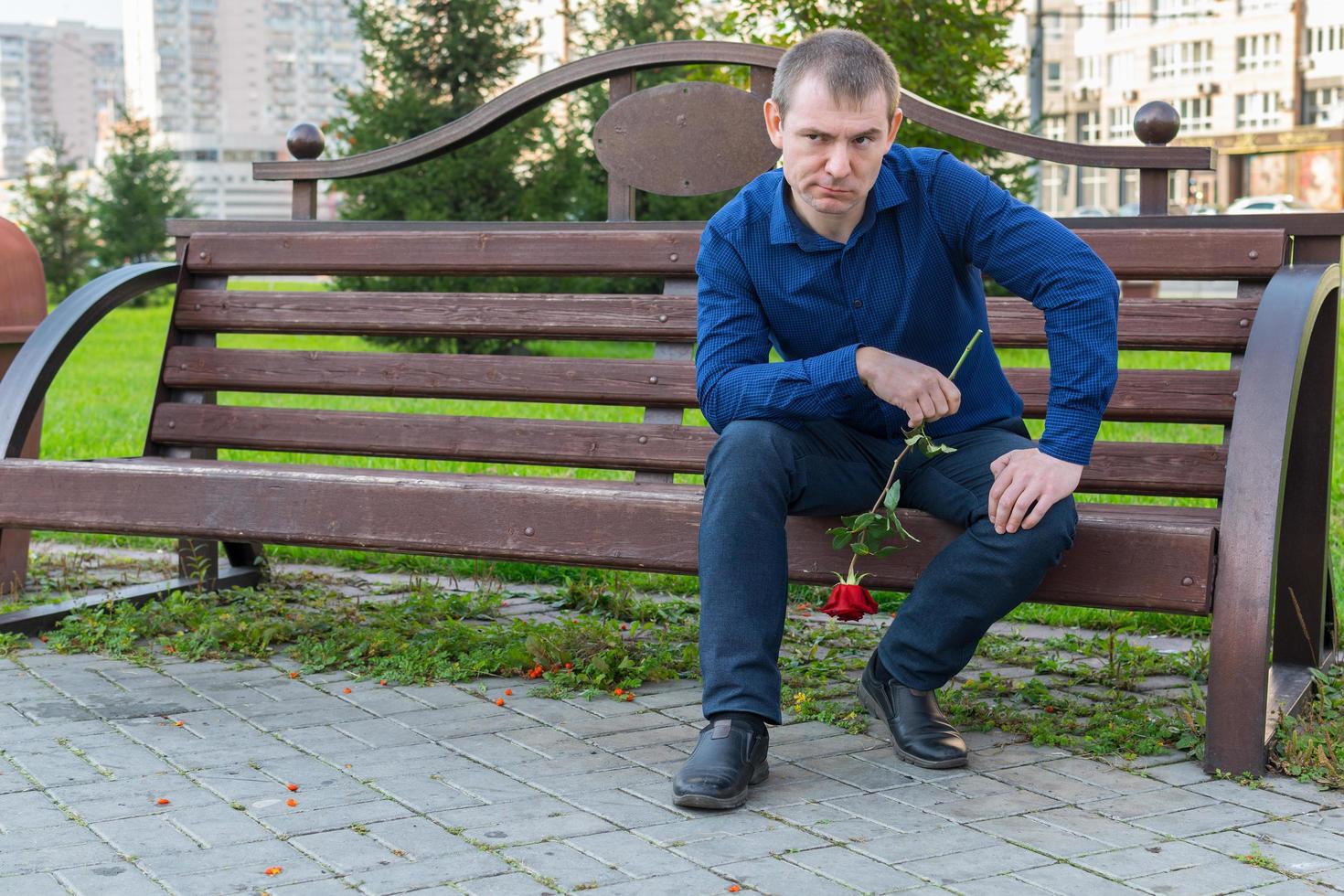 a man in a blue shirt and with a rose flower sits on a bench and examines the passing ladies photo