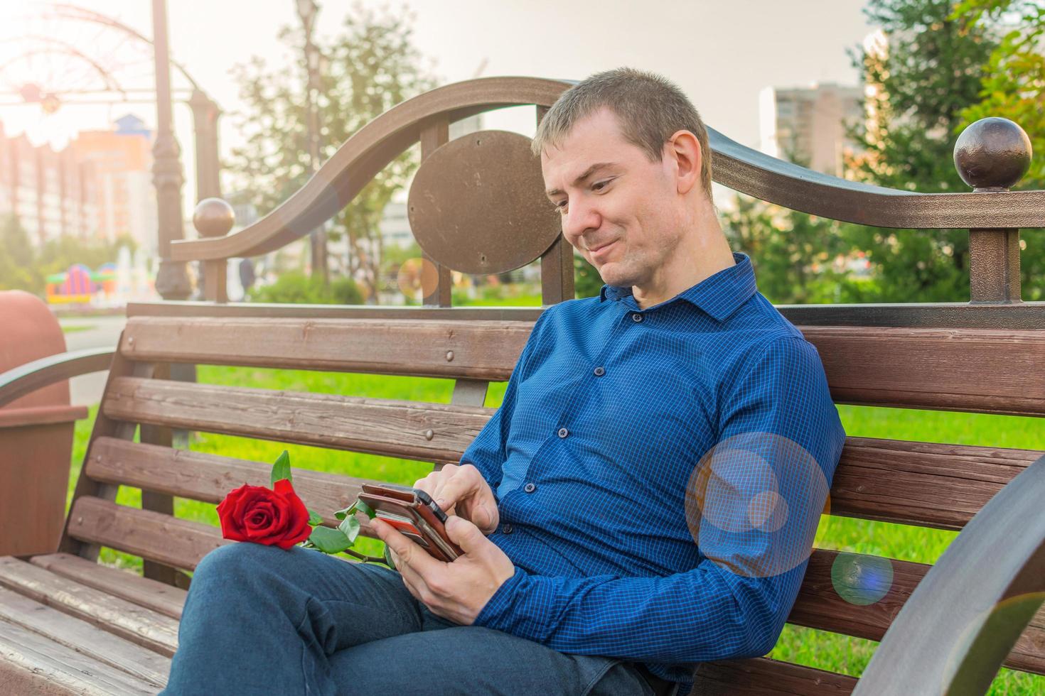 Happy man in a blue suit and with a red rose, sits on a park bench and carries on correspondence photo