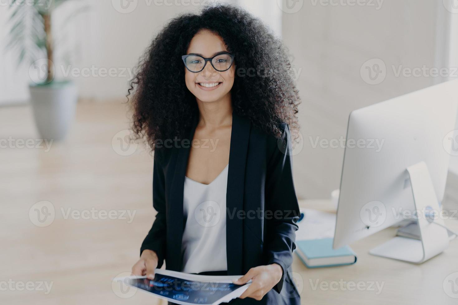 Pleased skilled dark skinned curly woman holds some papers, poses in modern office interior, wears transparent glasses and formal suit, stands near table with computer, studies documentation photo