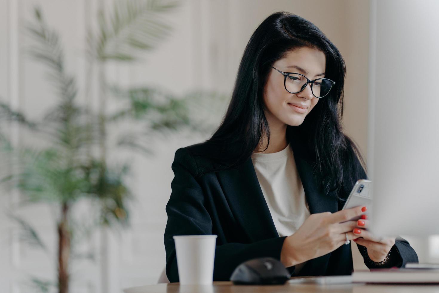 Satisfied business lady uses smartphone, poses at work place in front of computer screen, cup of tasty beverage, sends messages during break, poses at contemporary office. Technology and work concept photo