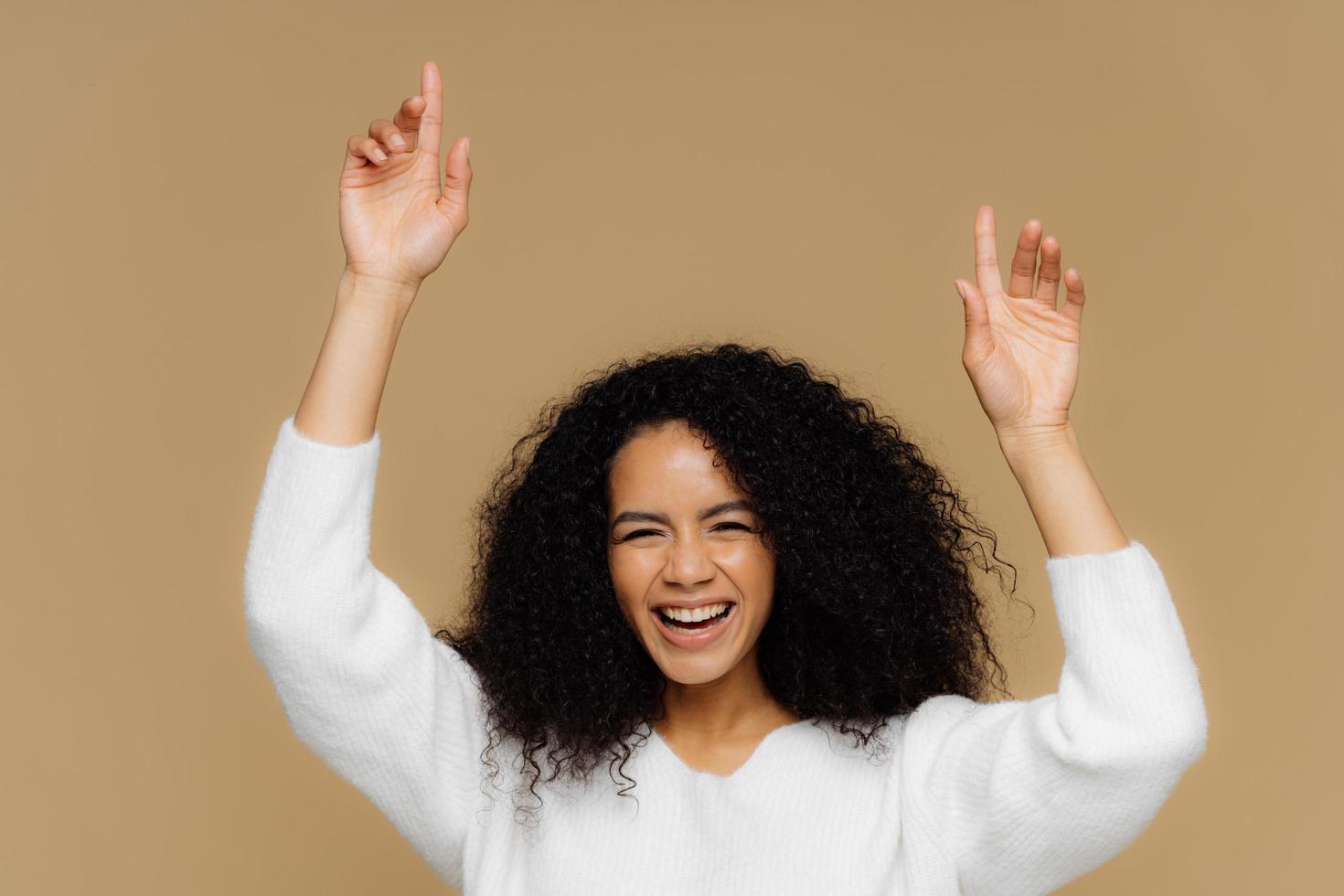 la foto horizontal de una encantadora modelo femenina de piel oscura tiene una sonrisa con dientes, levanta las manos, señala hacia arriba, se siente encantada, tiene el pelo rizado, usa un suéter blanco, aislada sobre un fondo marrón. la mujer se divierte