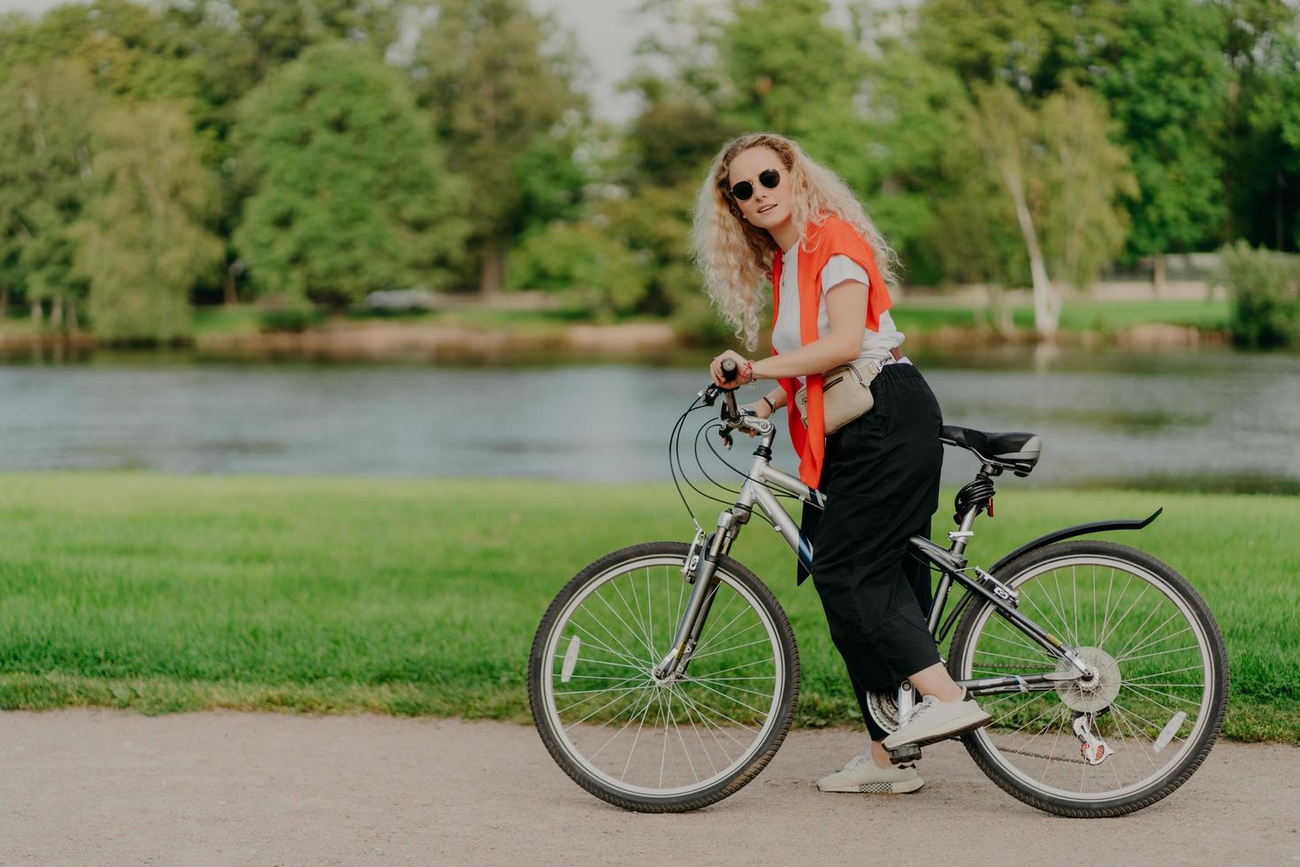 toma horizontal de una ciclista activa que usa camiseta, pantalones negros y zapatillas de deporte, se detiene cerca del lado del río, mantiene los pies en los pedales de la bicicleta hermosa naturaleza escénica en el fondo. día soleado, descanso activo foto