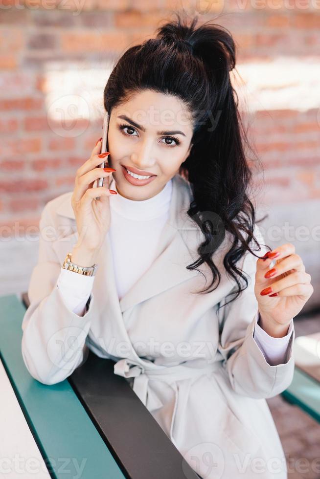 joven mujer carismática con cola de caballo ondulada oscura con manicura roja vestida elegantemente hablando en su teléfono móvil con una sonrisa mientras estaba sentada en el café esperando su pedido. concepto de comunicación foto