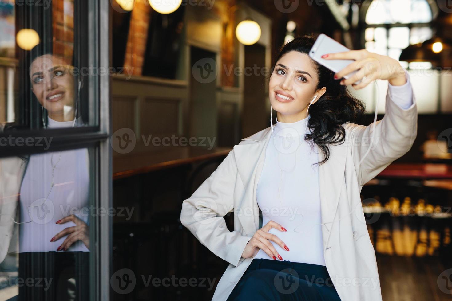 Waist up portrait of beautiful lady with dark hair tied in pony tail wearing white jacket sitting in cafe making selfie using her modern smartphone and listening to music with earphones. People, rest photo