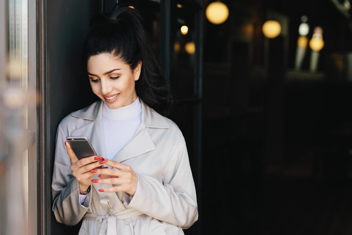 mujer hermosa de moda con cabello oscuro atado en cola de caballo vestida con un elegante abrigo blanco sosteniendo un teléfono celular mirando a la pantalla con una expresión encantadora. modelo joven y bonita comunicándose en línea foto