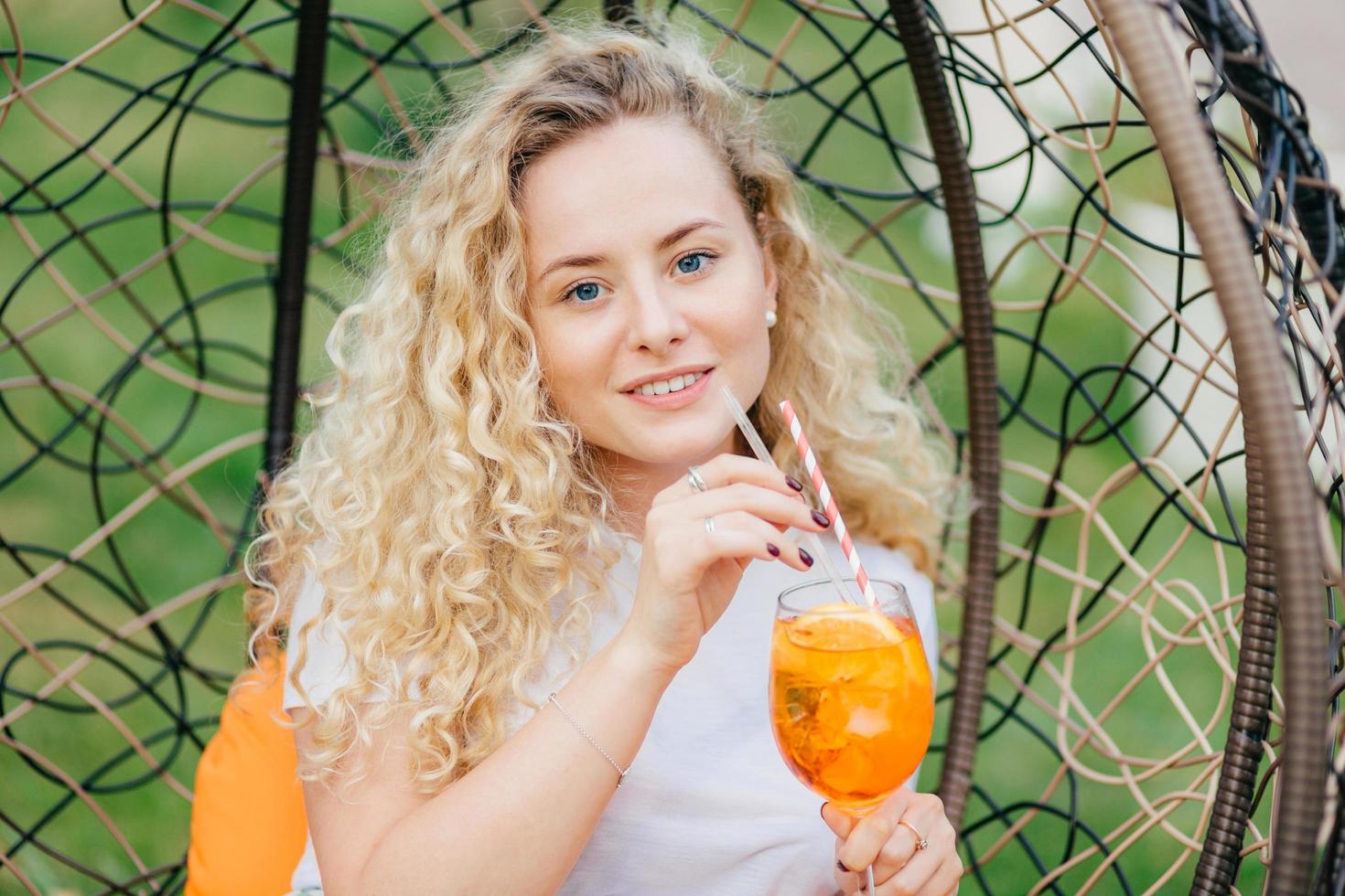 Outdoor shot of pleasant looking female with curly light hair, drinks fresh cold drink, rests in hanging chair, breathes fresh air, has summer party with friends. People and lifestyle concept. photo