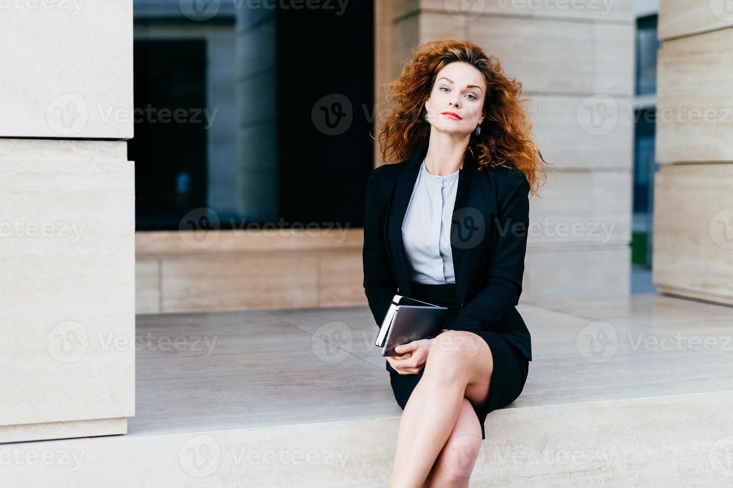 Adorable female model with bushy wavy hair, red lips and shining eyes, dressed formally while crossing her legs, holding tablet with pocket book, looking confidently into camera. Business concept photo