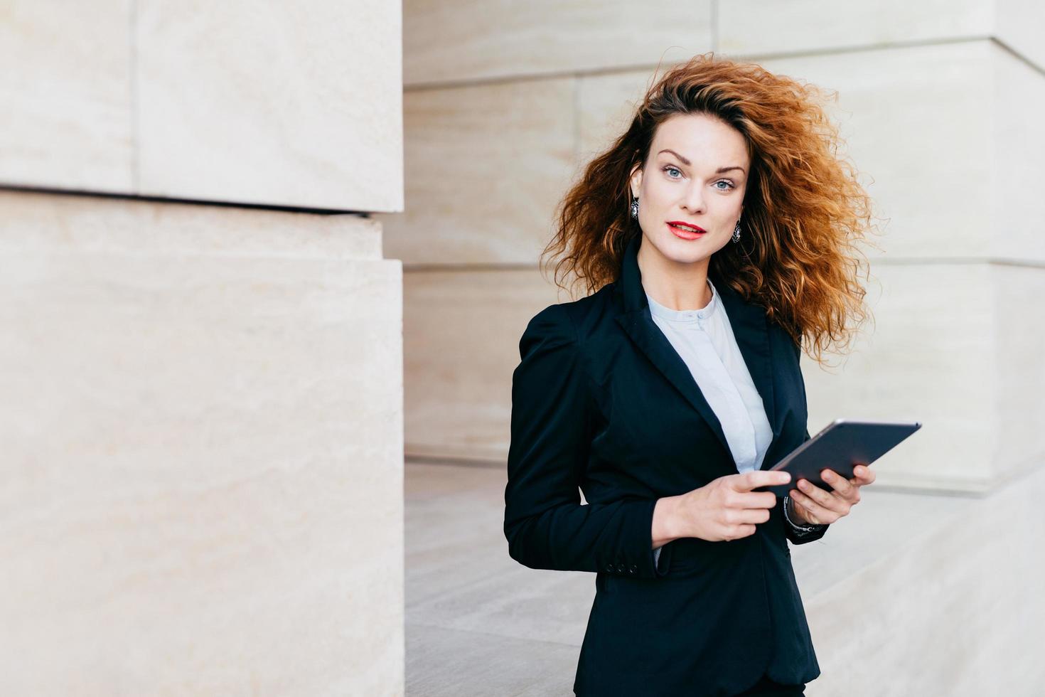 Beautiful businesswoman with curly fluffy hair, wearing elegant clothes, holding tablet in hands, waiting for her business partner near cafe, using free internet conncection. People, career, work photo