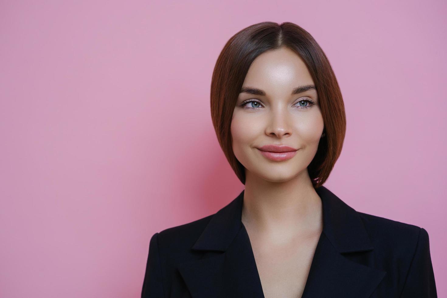 hermosa mujer de negocios mira a un lado con expresión soñadora, piensa en algo agradable durante el descanso después del trabajo, usa ropa negra, aislada en un fondo rosa, espacio vacío para su información foto