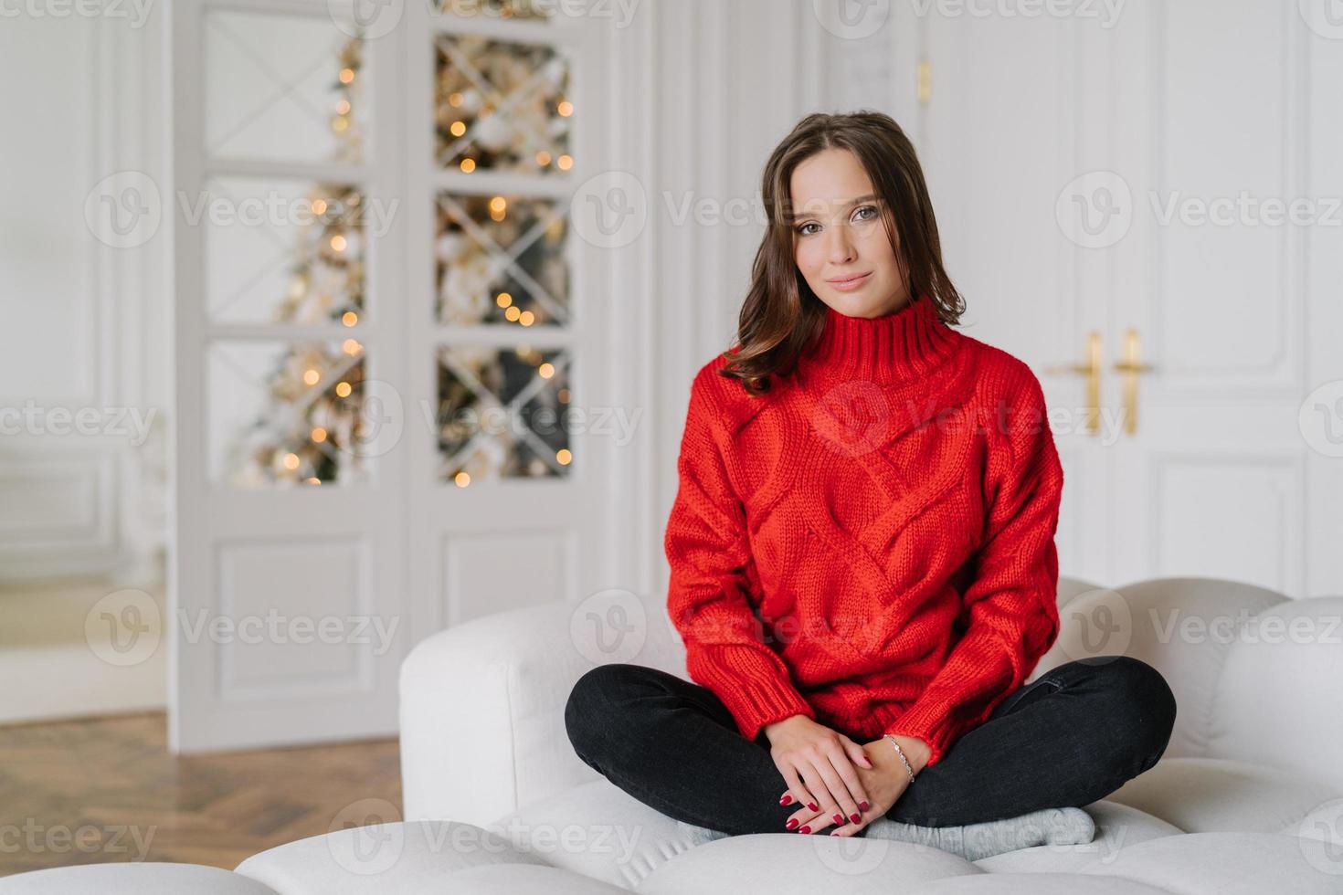 Horizontal shot of relaxed housewife dressed in warm red knitted sweater, sits crossed legs in lotus pose on couch, in spaciouss room with New Year tree in background, light colours. Rest concept photo