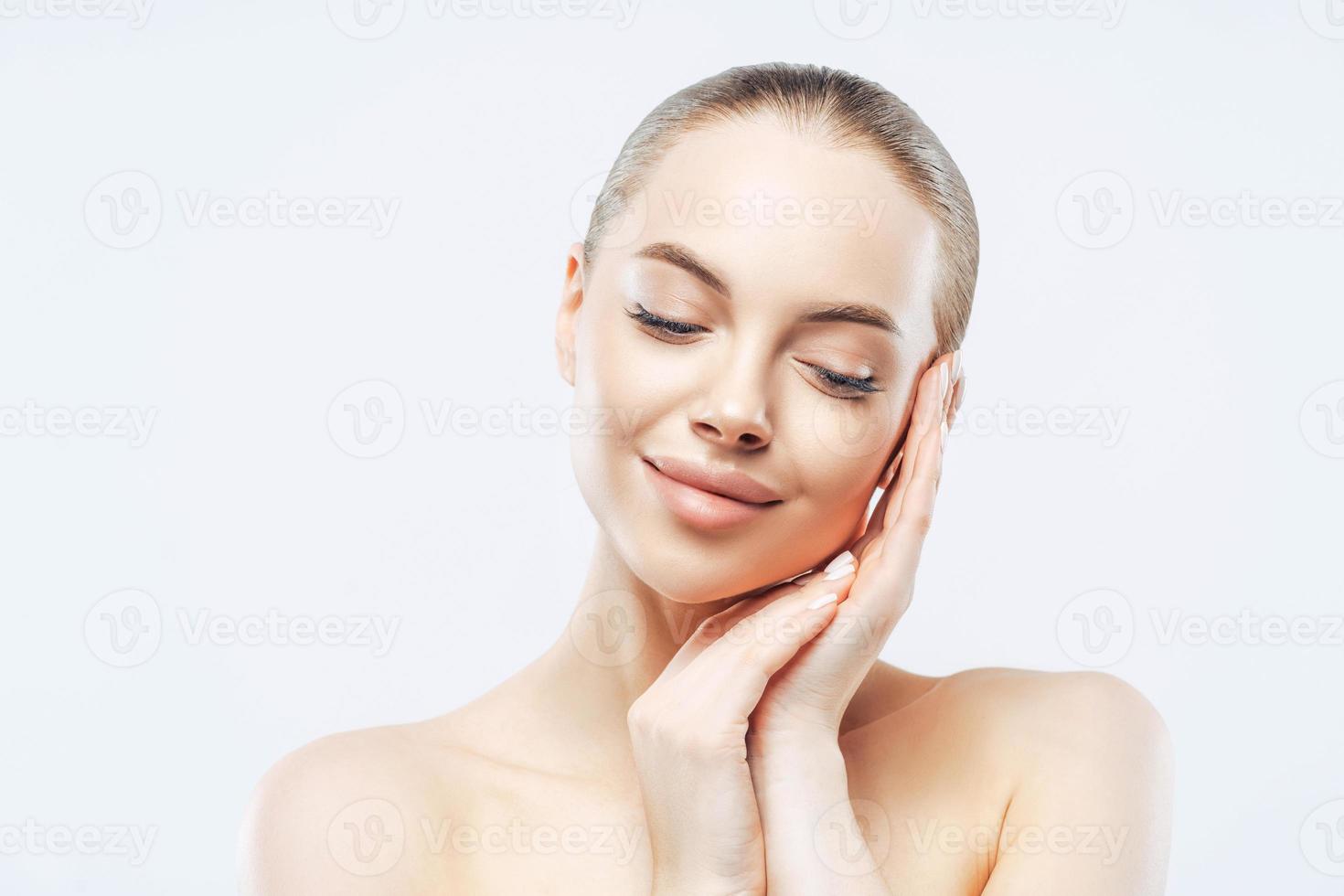 Headshot of tender beautiful woman touches cheek and stands naked indoor, enjoys daily hygiene procedure, looks down, cares about skin, being in romantic mood, stands over white studio wall. photo