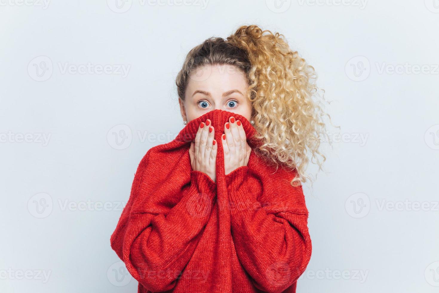 Frightened stunned female sees her phobia, covers face with collar of red sweater, stares at camera with scared look, isolated over white studio background. People, reaction and emotions concept photo