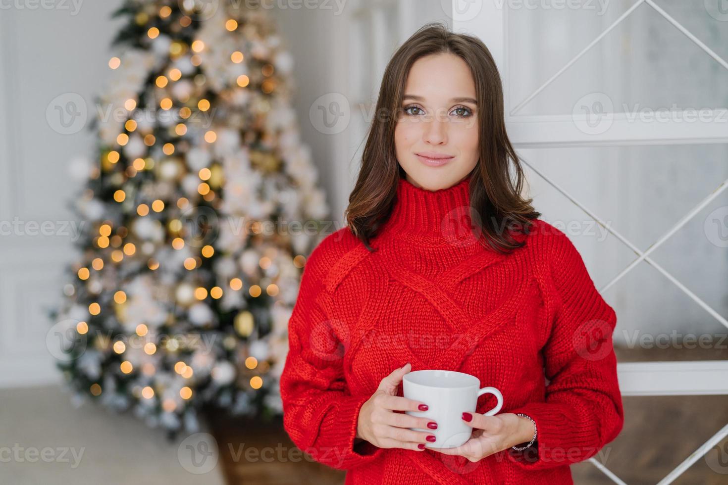 toma interior de una hermosa mujer caucásica vestida con un suéter de guerra, bebe bebidas calientes, disfruta del ambiente doméstico, se prepara para celebrar la navidad o el año nuevo. concepto de personas y estilo de vida foto