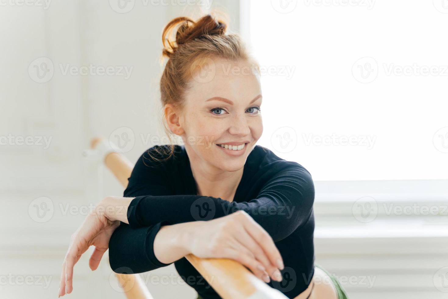 Fitness female with red combed hair, wears special clothes, practices ballet on barre in dancing studio, has toothy smile, being in high spirit, wears makeup, has healthy lifestyle. Training concept photo