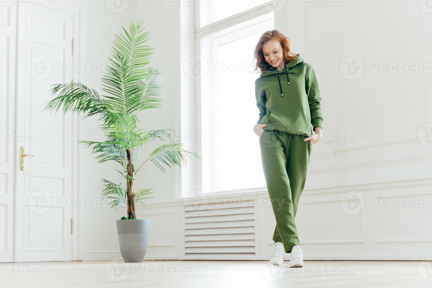Full length shot of active pleased young woman with slim body, keeps hands in pockets of tracksuit, focused down, ready for doing cardio exercises, has determined look, pilates exercising indoor photo
