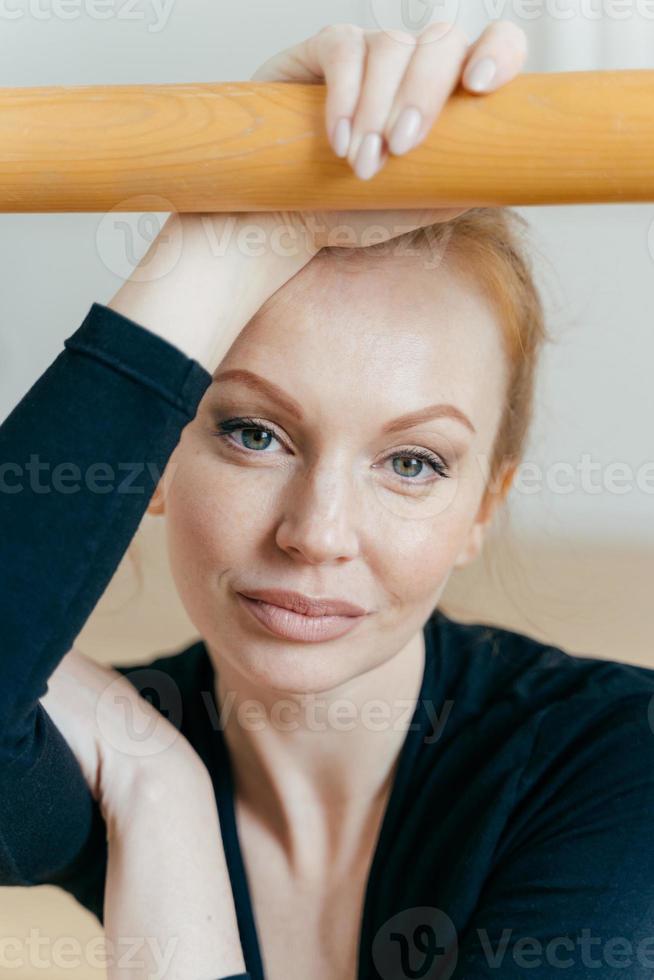Close up shot of restful woman with ginger hair, has make up, looks at camera, has serious expression, holds hand on handrail, busy doing gymnastic exercises, has natural beauty. Ballet concept photo