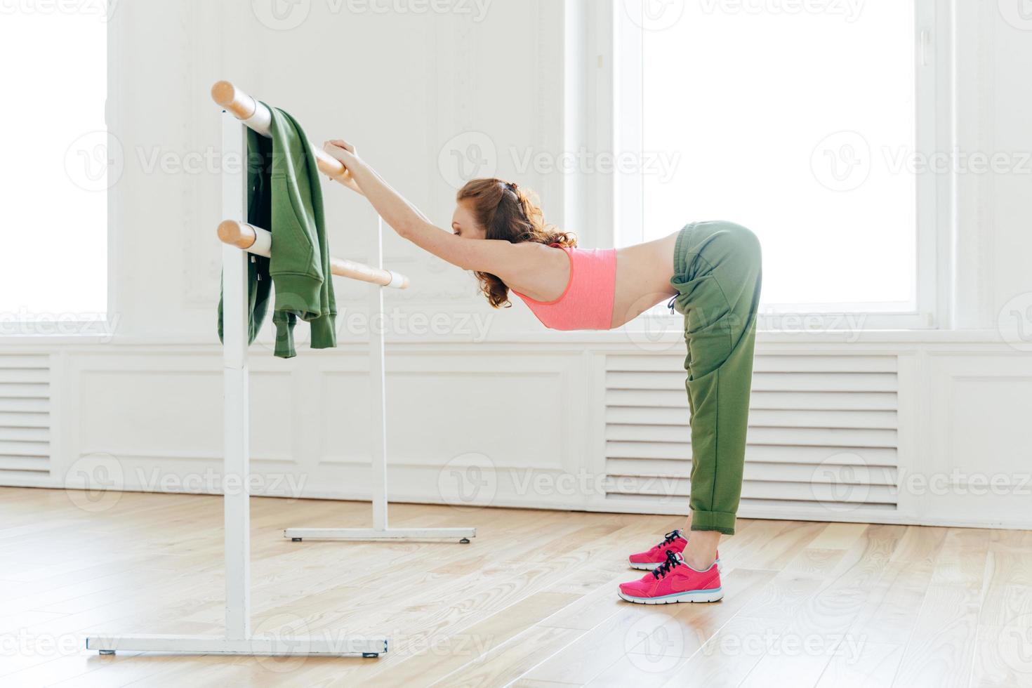 Indoor shot of sportswoman in sportsbra and active wear, leans at ballet barre, has productive training session, slim body shape, stretches muscles, has motivation for being fit and healthy. photo