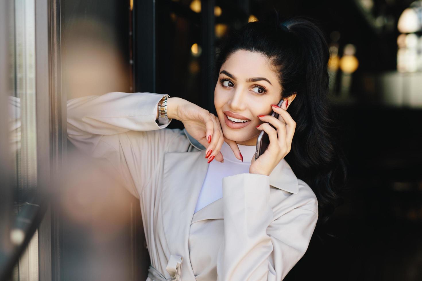 mujer atractiva con cabello oscuro y ojos agradables y atractivos con labios carnosos vestidos con ropa elegante conversando con su mejor amiga por teléfono celular mirando a un lado. gente, concepto de belleza foto