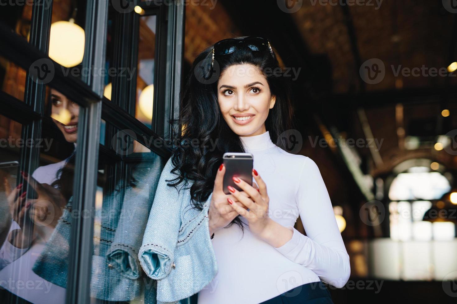 mujer atractiva con cabello oscuro, ojos brillantes y pestañas largas con gafas de sol en la cabeza mirando pensativamente hacia arriba teniendo algunos sueños mientras se comunica con su amante a través de las redes sociales, usando internet foto