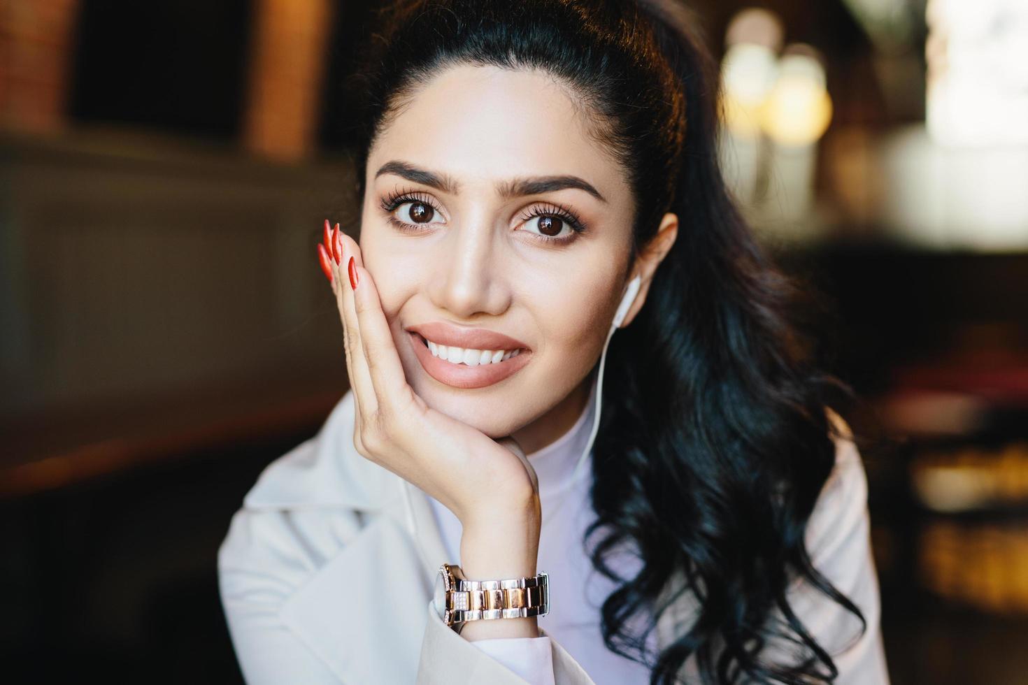 Close up portrait of beautiful brunette female with dark warm eyes, curling eyelashes, thin well-shaped eyebrows and full lips holding hand on cheek wearing elegant watch having earphones in ears photo