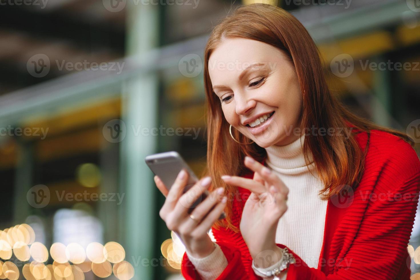 Positive pleased female model uses mobile phone data, uses chat application, holds modern cell phone, smiles positively, dressed in red coat, waits for answer or call from boyfriend, has brown hair photo