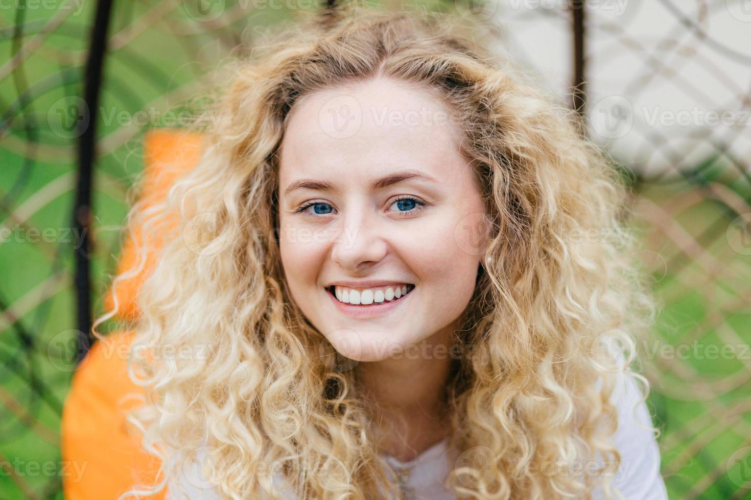 Photo of attractive curly blonde female with shining smile, shows white even teeth, being in high spirit, enjoys good rest outdoor, talks with friend. European woman poses in hanging chair outside