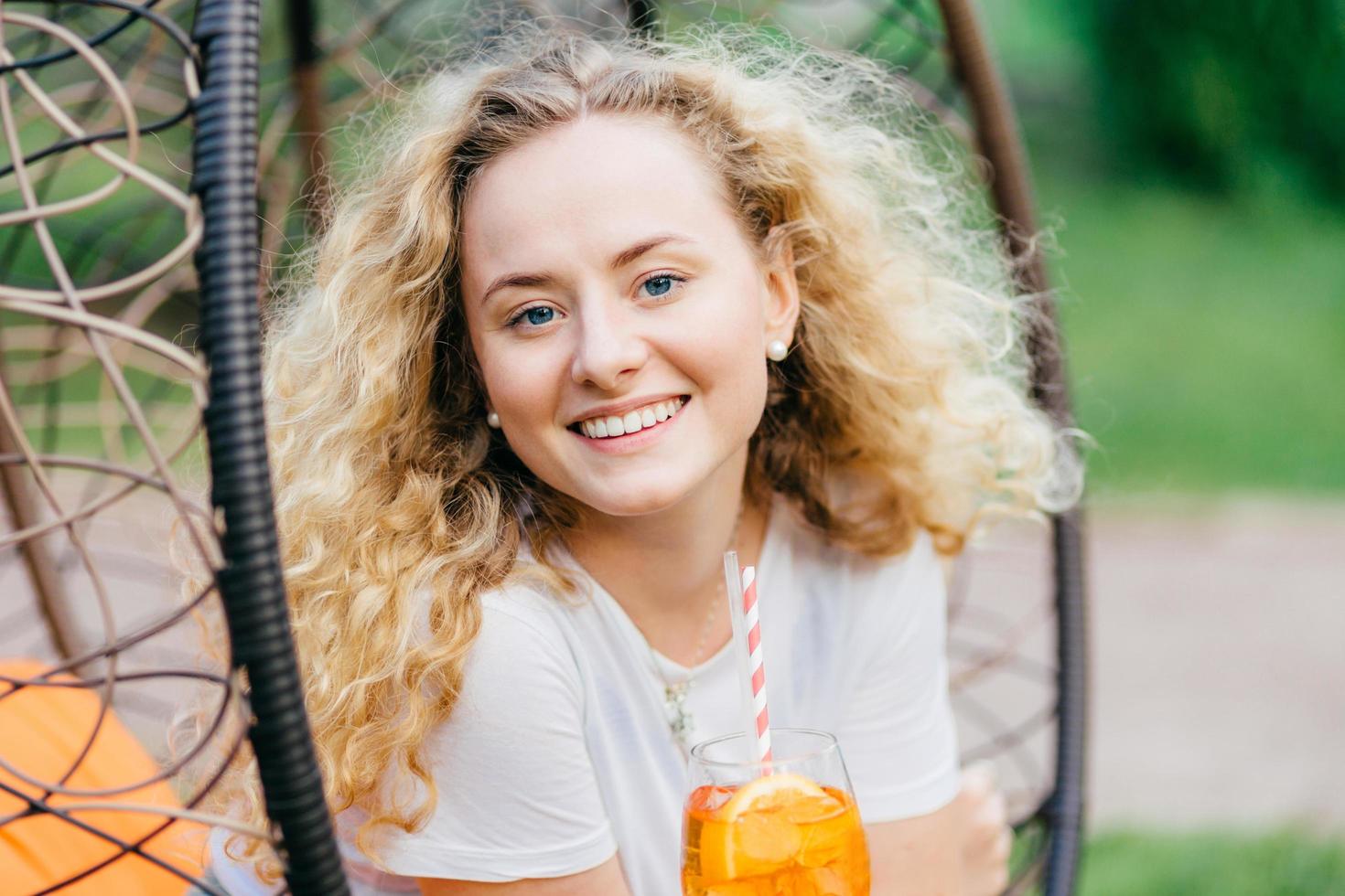 Horizontal shot of pretty smiling European female has wavy bushy light hair, holds fresh cocktail, sits on hanging chair, breathes fresh air. Positive emotions, recreation and people concept photo