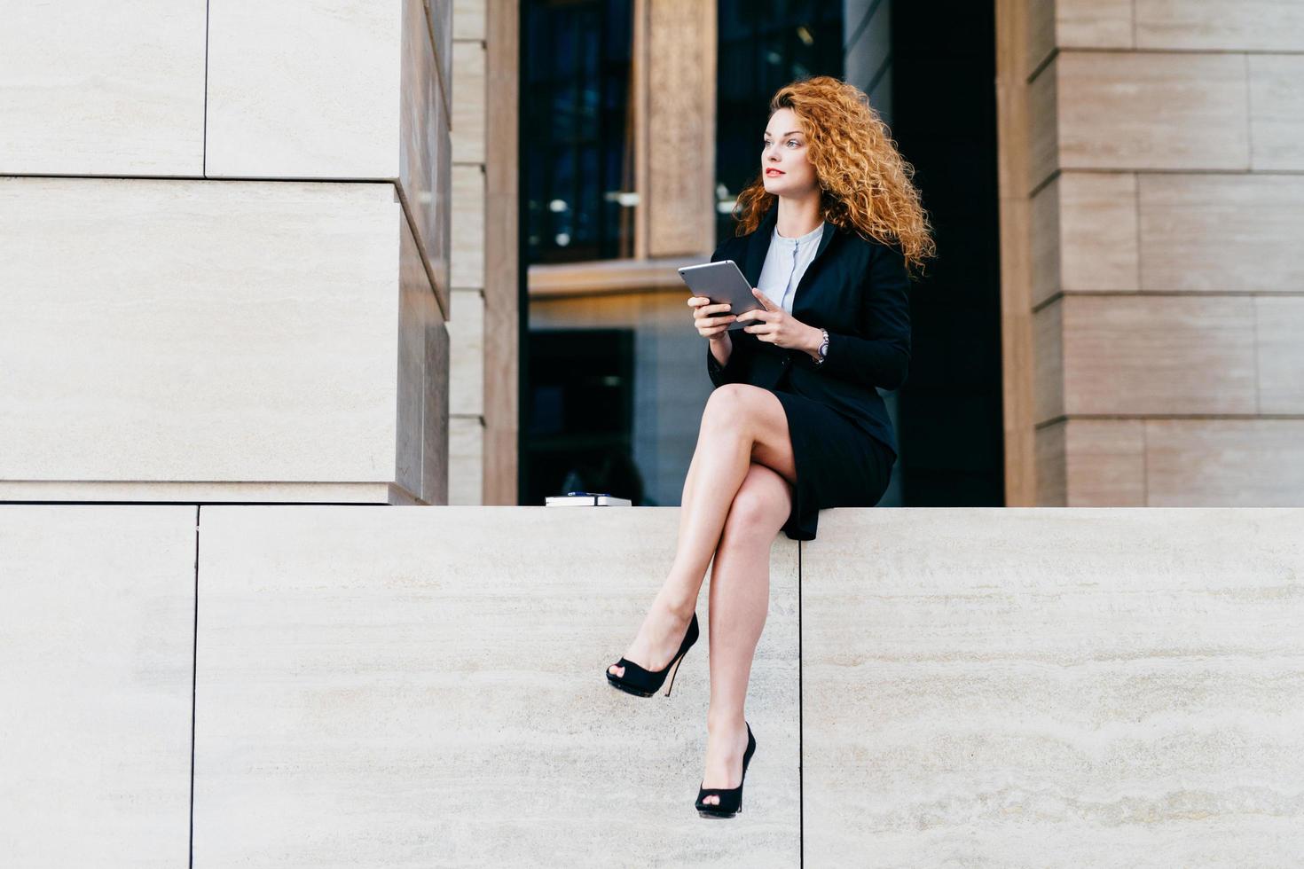 Slim businesswoman with wavy luxurious hair, having slender legs, wearing black elegant suit and shoes, holding tablet computer, looking aside while resting for minute after hard work in office photo