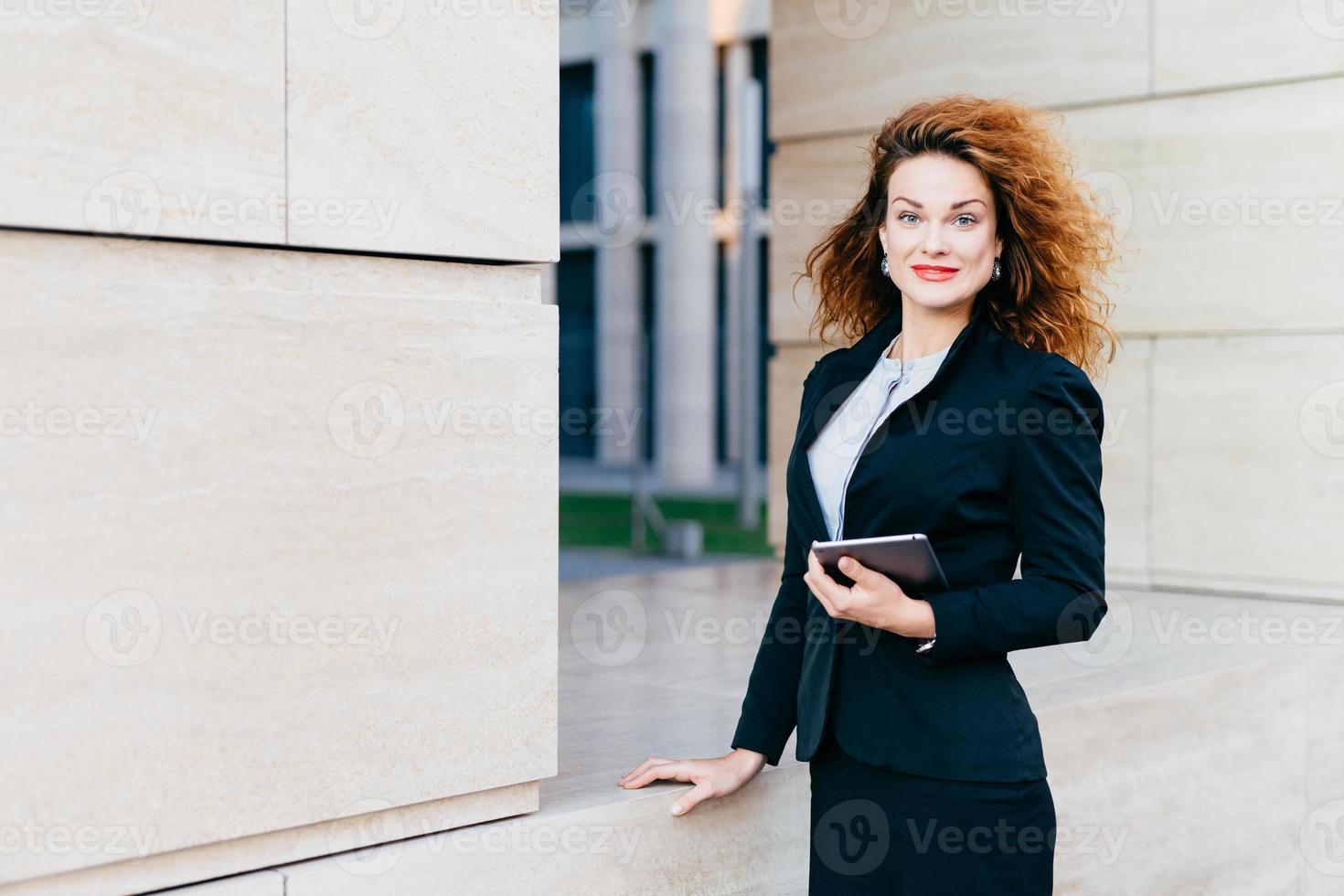 linda mujer independiente con lujoso cabello claro, ojos azules, labios rojos y piel pura, vestida con traje formal negro, con un aparato electrónico y las manos, reuniéndose con sus socios comerciales foto