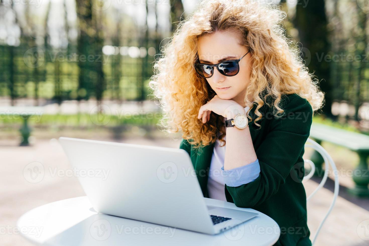 Young woman with blond curly hair wearing sunglasses and elegant green jacket sitting in front of open laptop outdoors reading news online having pensive expression. People, technology concept photo