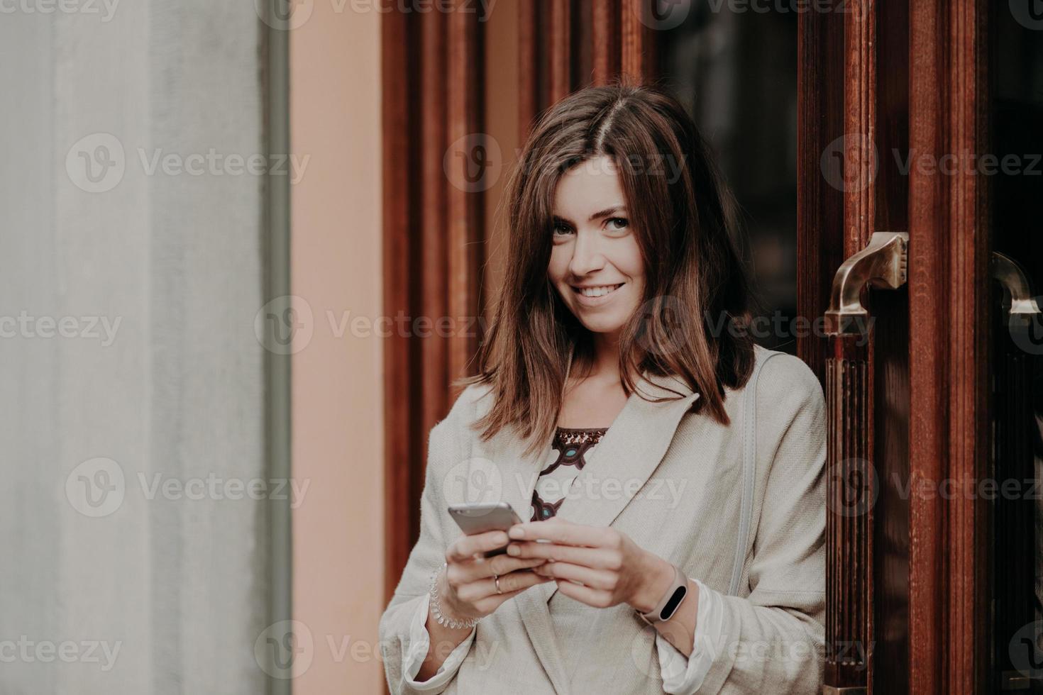 Brunette cheerful lovely woman texts email letter, uses mobile phone and free internet connection, dressed in white jacket, stands near doors of building. People, lifestyle, technology concept photo