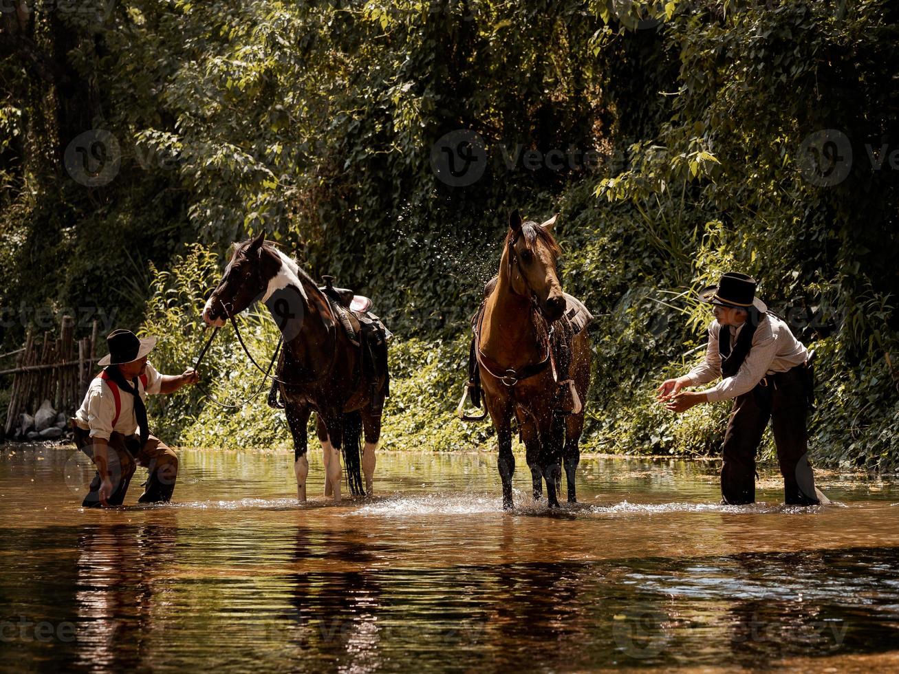 Senior and young cowboys stand to bathe their horses in a forest stream photo