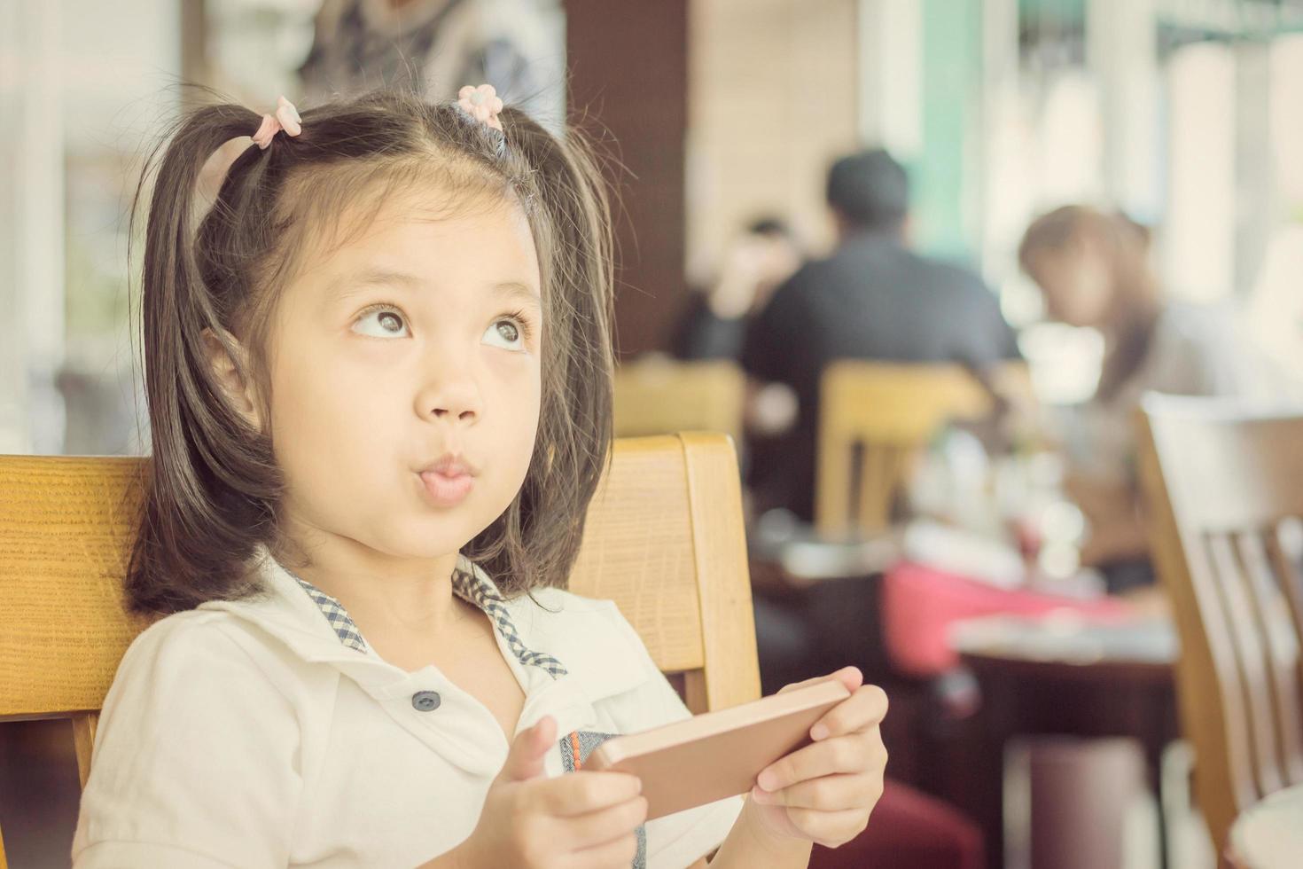 primer plano de una hermosa niña linda mirando hacia arriba pensando y sosteniendo un teléfono inteligente, concepto de niño feliz. foto