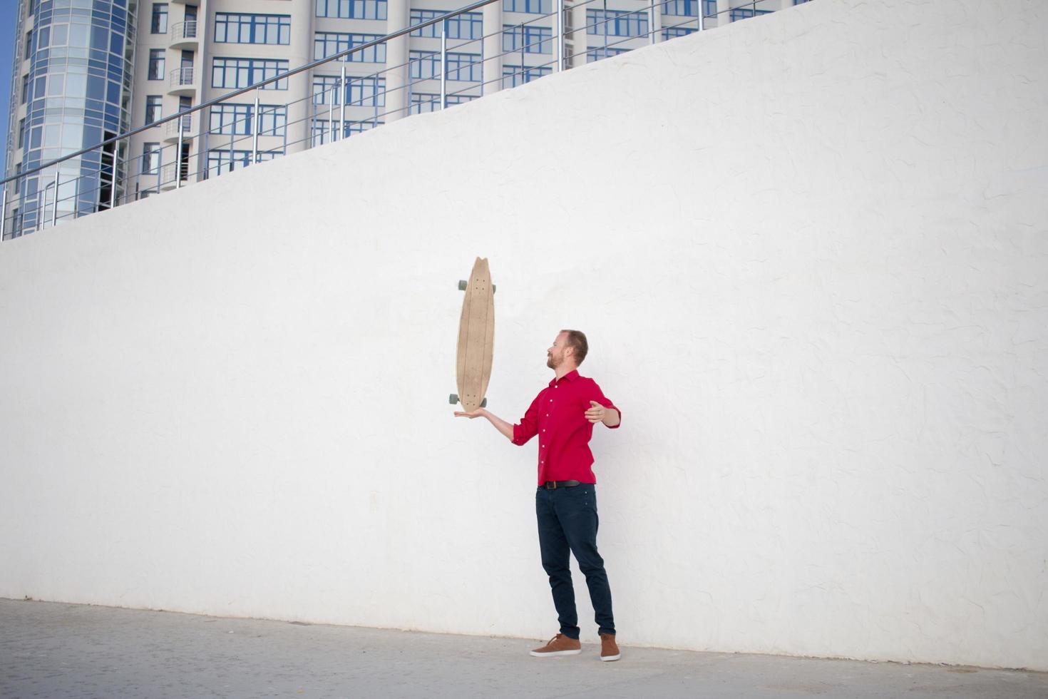 joven barbudo montando en patineta, hipster con longboard en camisa roja y blue jeans de fondo urbano foto