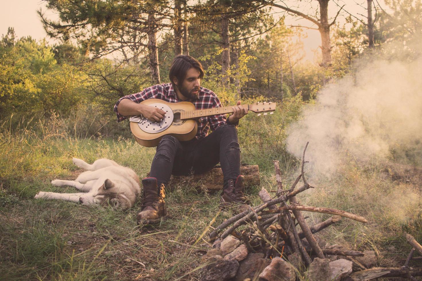 Folk or country guitar player in the forest with dog, man in woods, campfire and sunset background photo