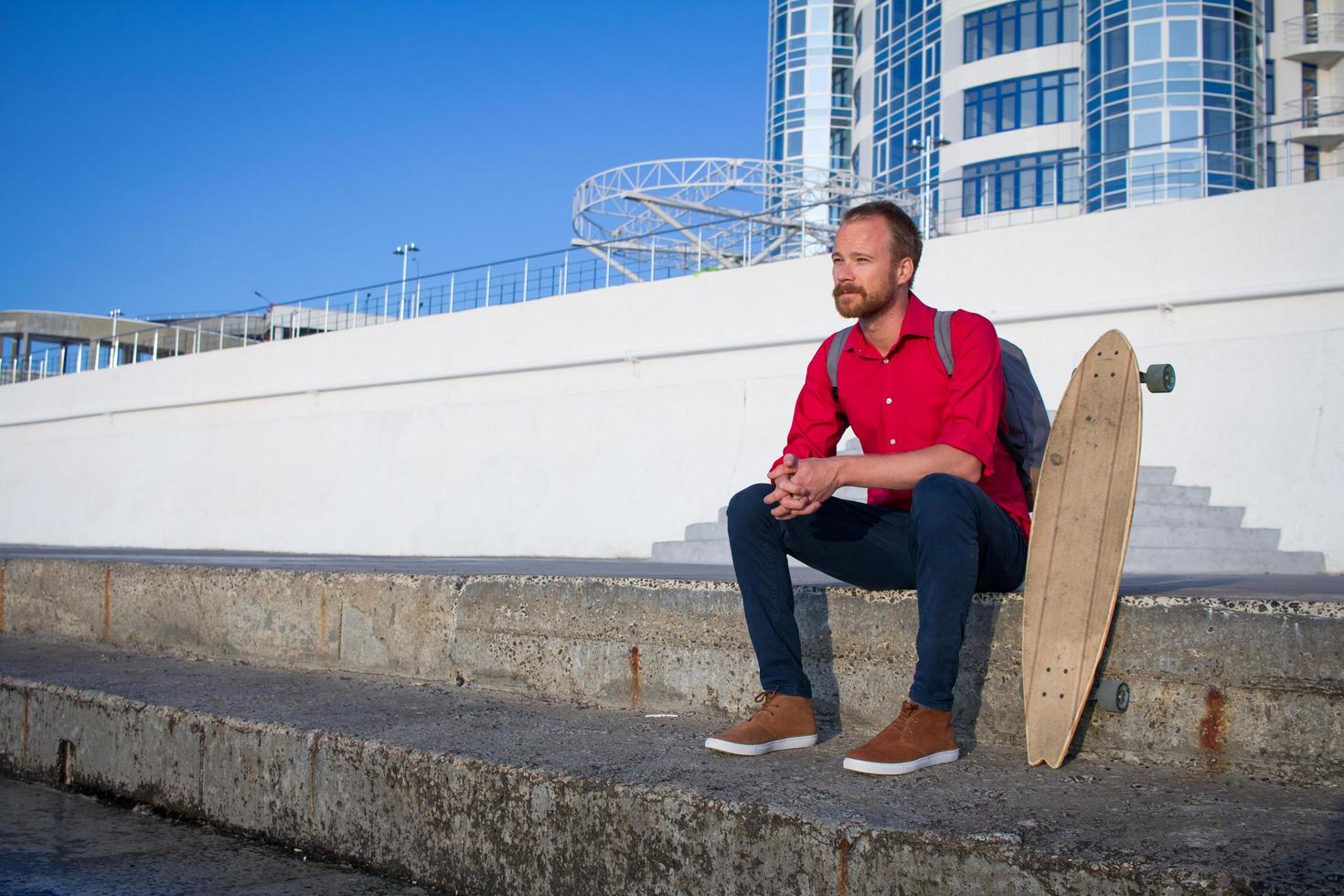 Young bearded man riding on skateboard, hipster with longboard in red shirt and blue jeans urban background photo