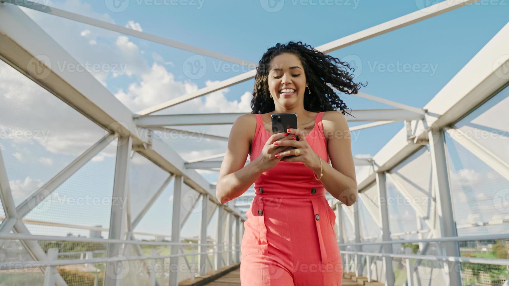 joven mujer latina de pelo rizado caminando con teléfono celular. enviando mensajes de texto en la calle. Gran ciudad. foto