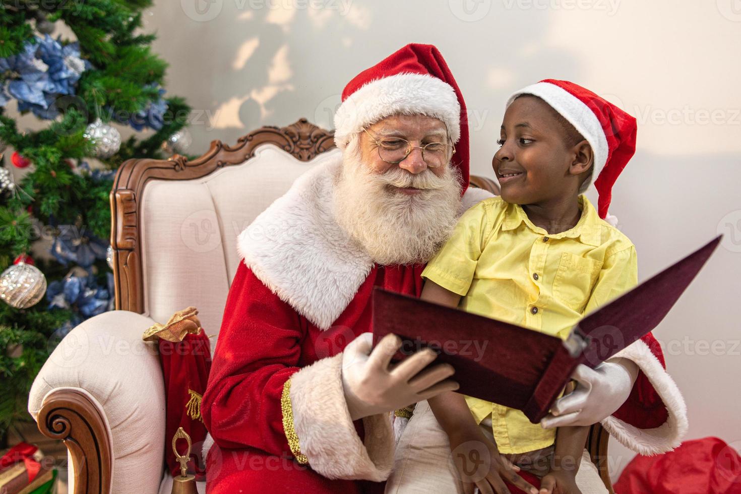 Santa Claus reading a storybook to a child on his lap. Education, beliefs and legends concept. Child delighted with what he learns. End of the year. Enchanted Fable. African boy. photo