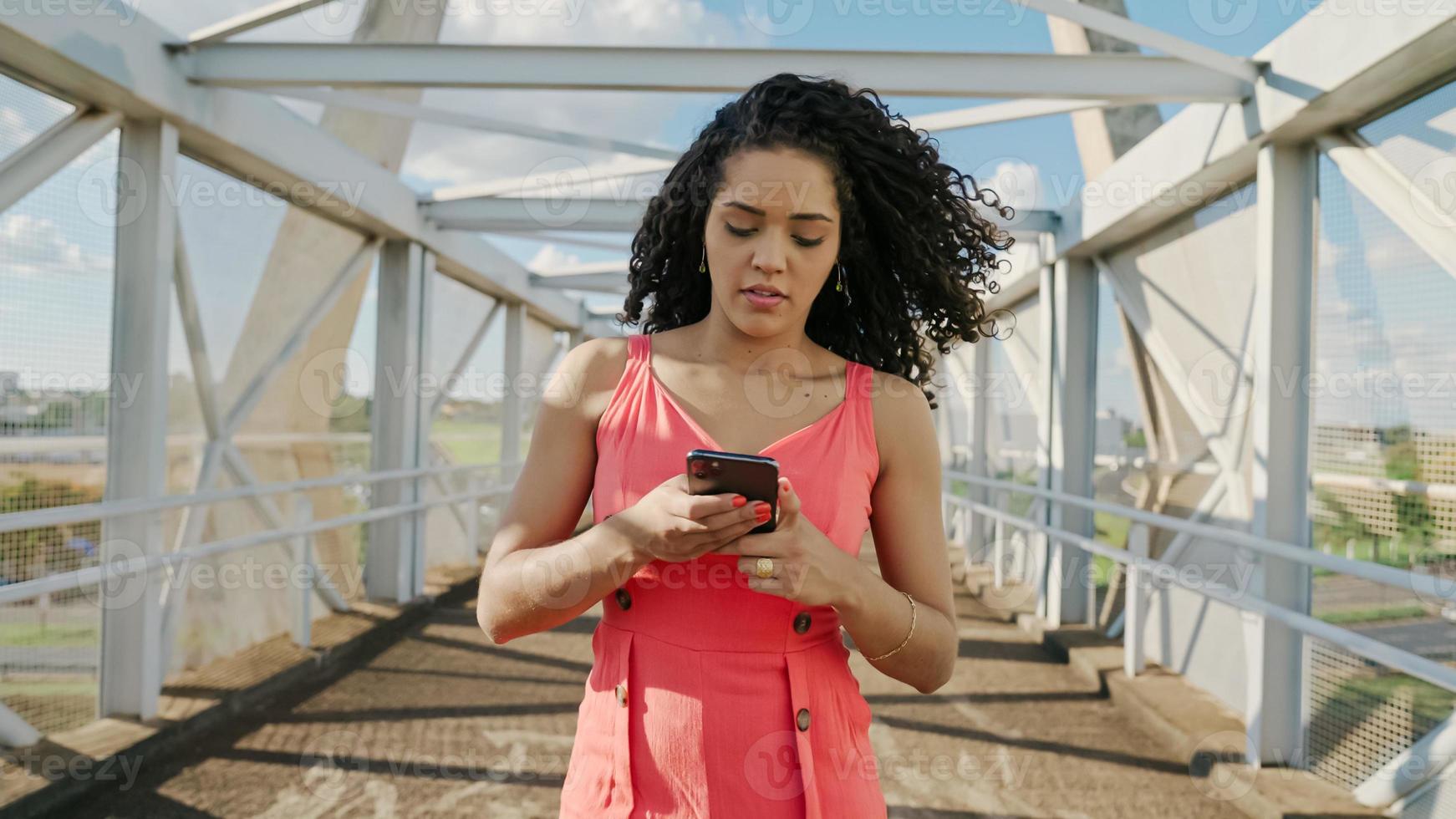 joven mujer latina de pelo rizado caminando con teléfono celular. enviando mensajes de texto en la calle. Gran ciudad. foto