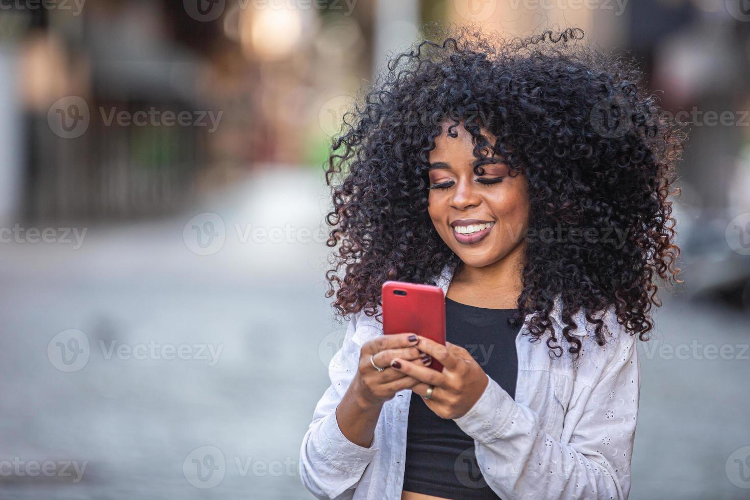 Young curly hair black woman walking using cell phone. Texting on street. Big city. photo