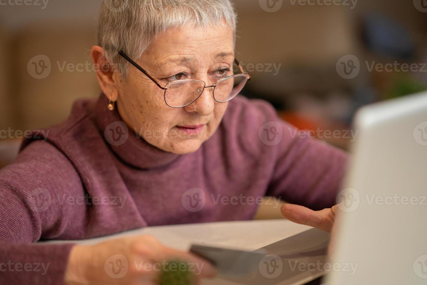 una mujer mayor con cabello gris se sienta en una computadora portátil con una tarjeta de crédito en las manos, hace compras en línea. concepto de compras en línea foto