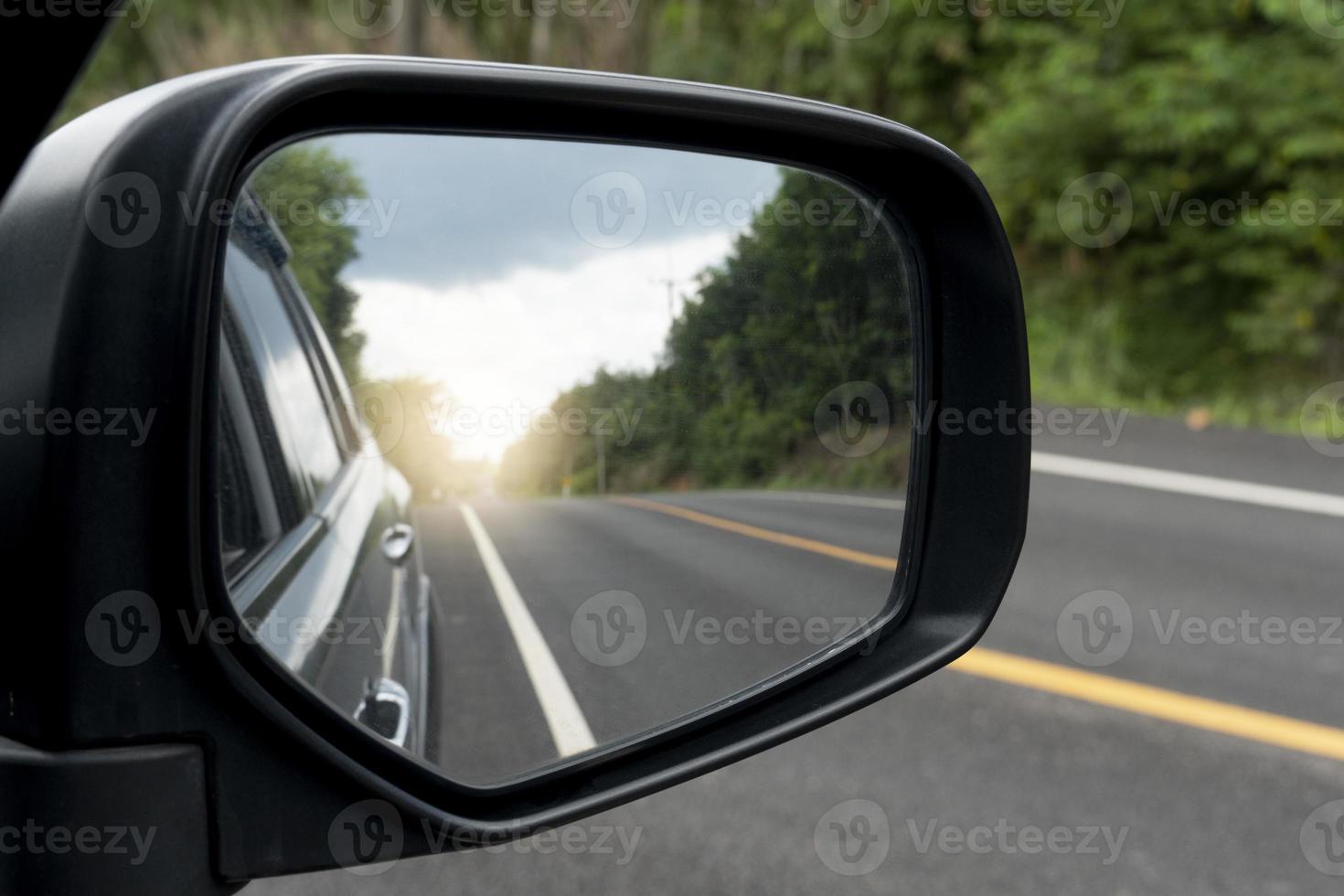 Inside view of mirrors wing. Rear view of a gray car with asphalt road and green trees in the daytime. Clear traffic in rural areas. photo