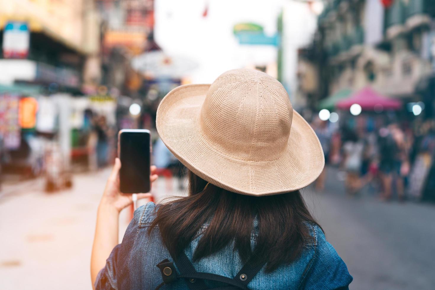 vista trasera de una joven viajera asiática de estilo casual usando un teléfono inteligente. foto