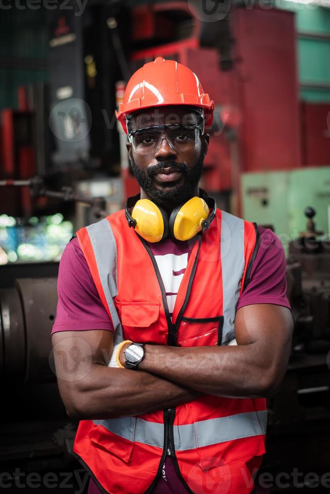 retrato de ojos sobrios y decididos trabajador afroamericano tiene barba descuidada en uniforme de seguridad con casco, gafas, chaleco y guante en la fábrica industrial. foto