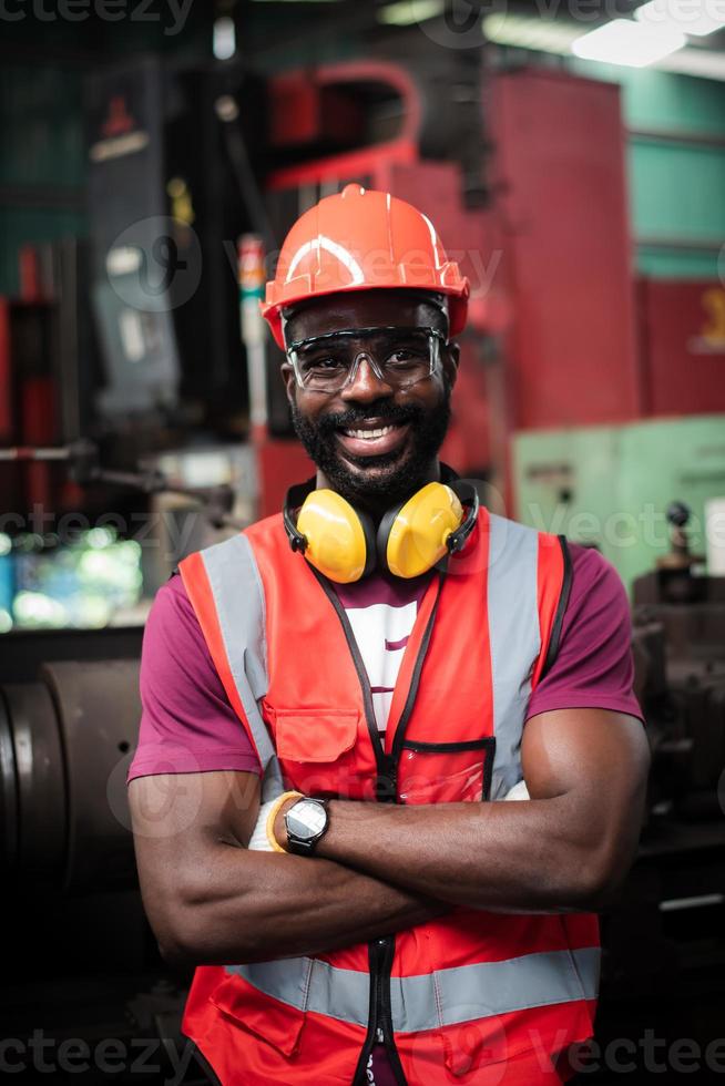 el retrato de un trabajador afroamericano feliz tiene barba descuidada con uniforme de seguridad usando casco, anteojos, chaleco y guante en una fábrica industrial. foto