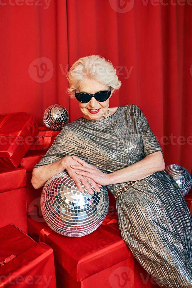 Senior gray haired woman in sunglasses and silver dress with disco ball at the party on red curtains background. party, celebration, senior age concept photo